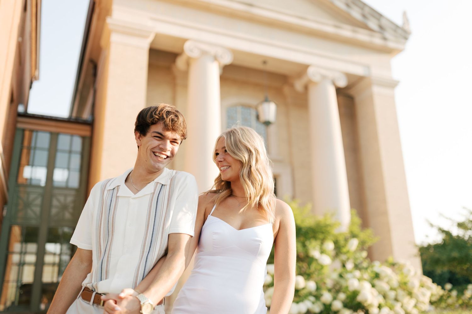 Couple in white outfits pose together against classical architecture.