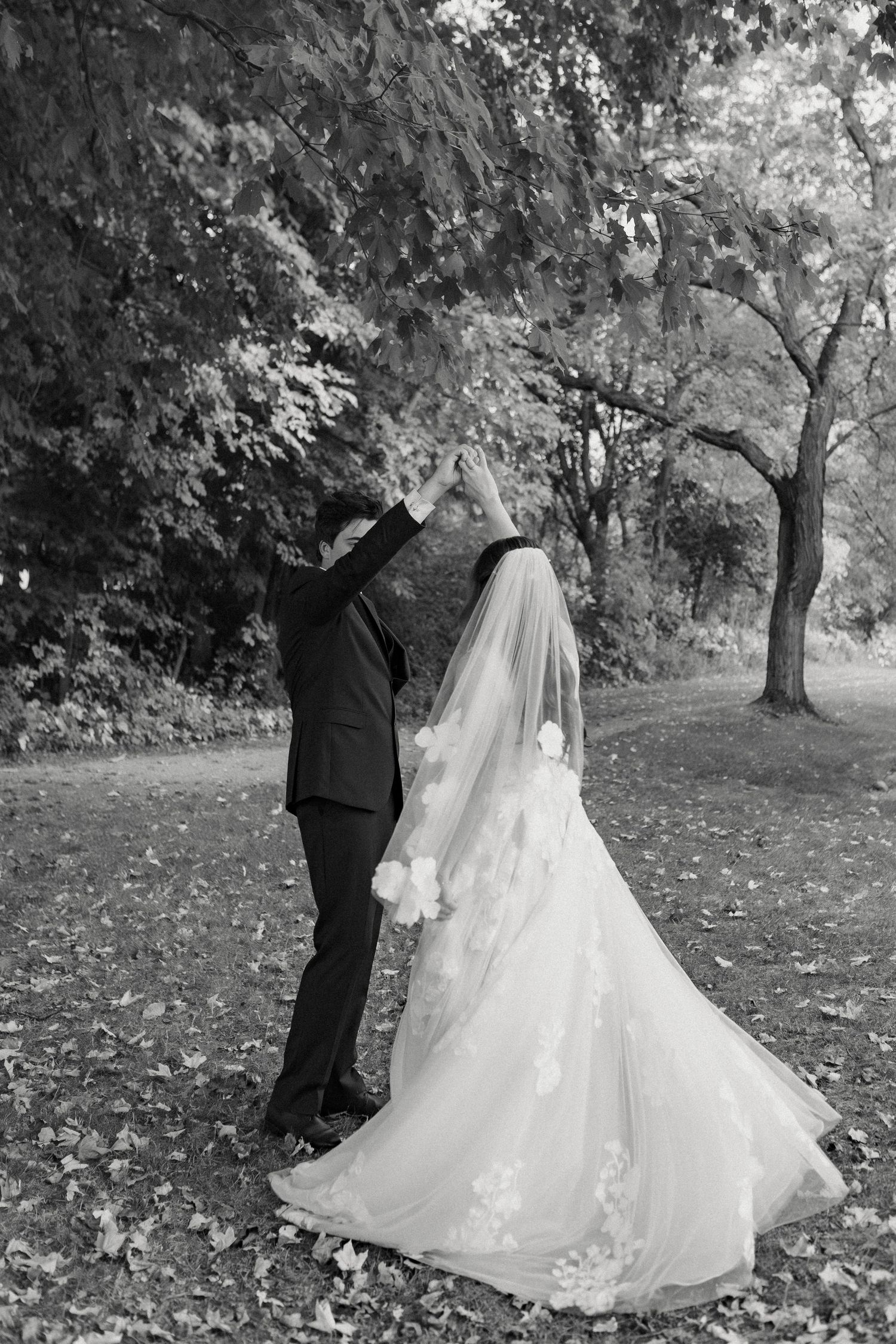 A couple in formal wedding attire dancing together in a scenic wooded outdoor setting with autumn trees.