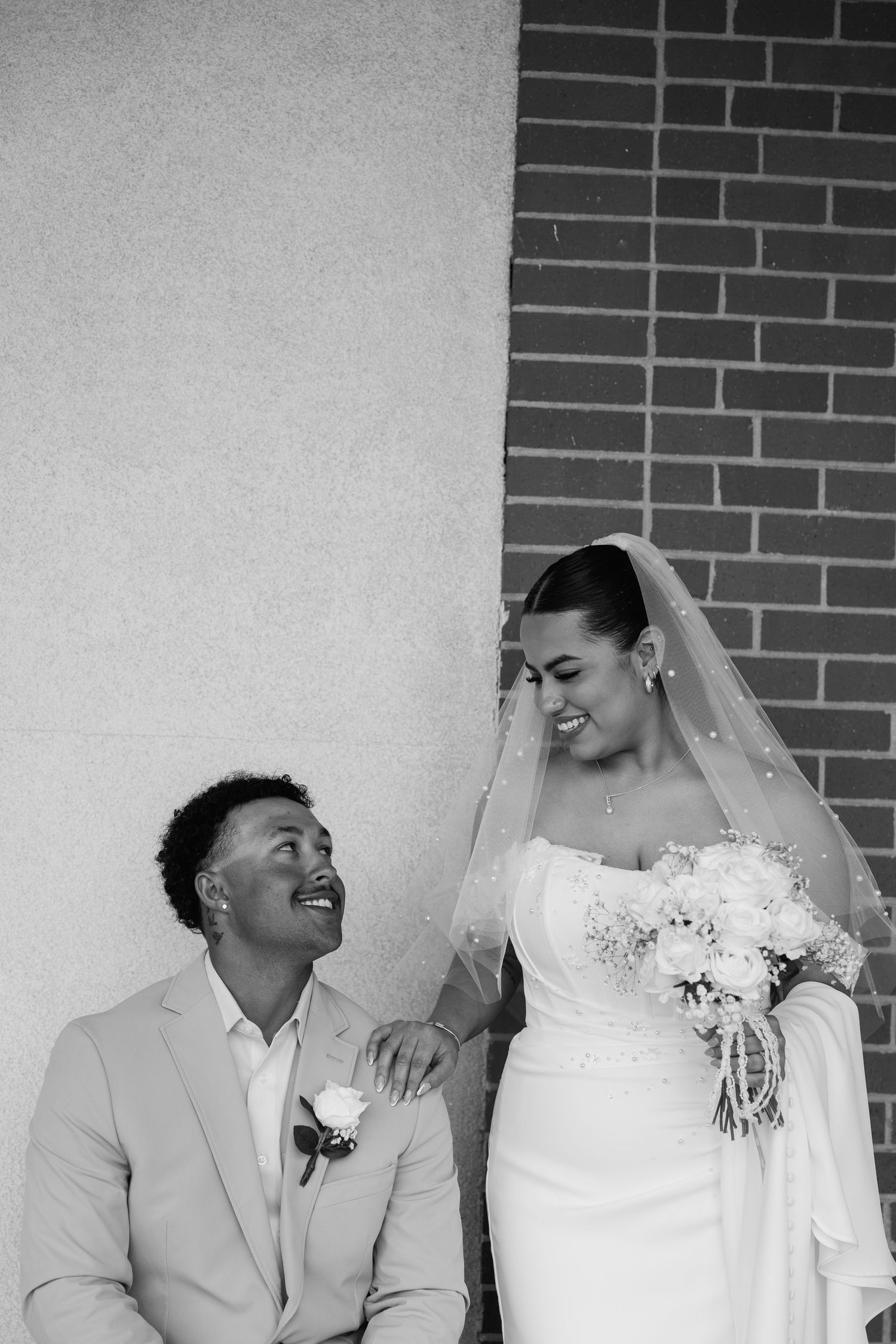 Black and white photo of a laughing groom in a light suit and bride in a wedding dress with veil holding a bouquet.