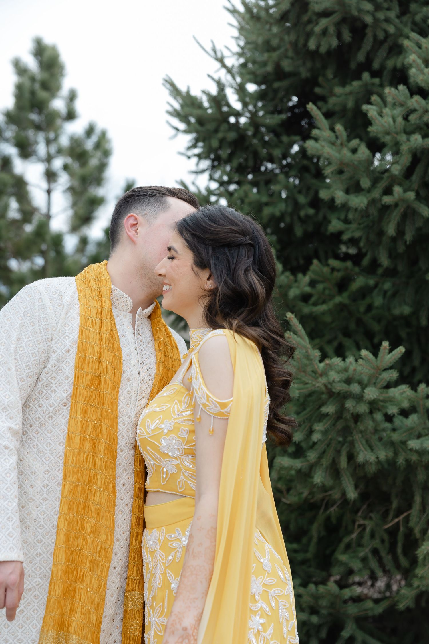 Couple in traditional Indian wedding attire, yellow lehenga and sherwani, smiling together outdoors near evergreen trees.