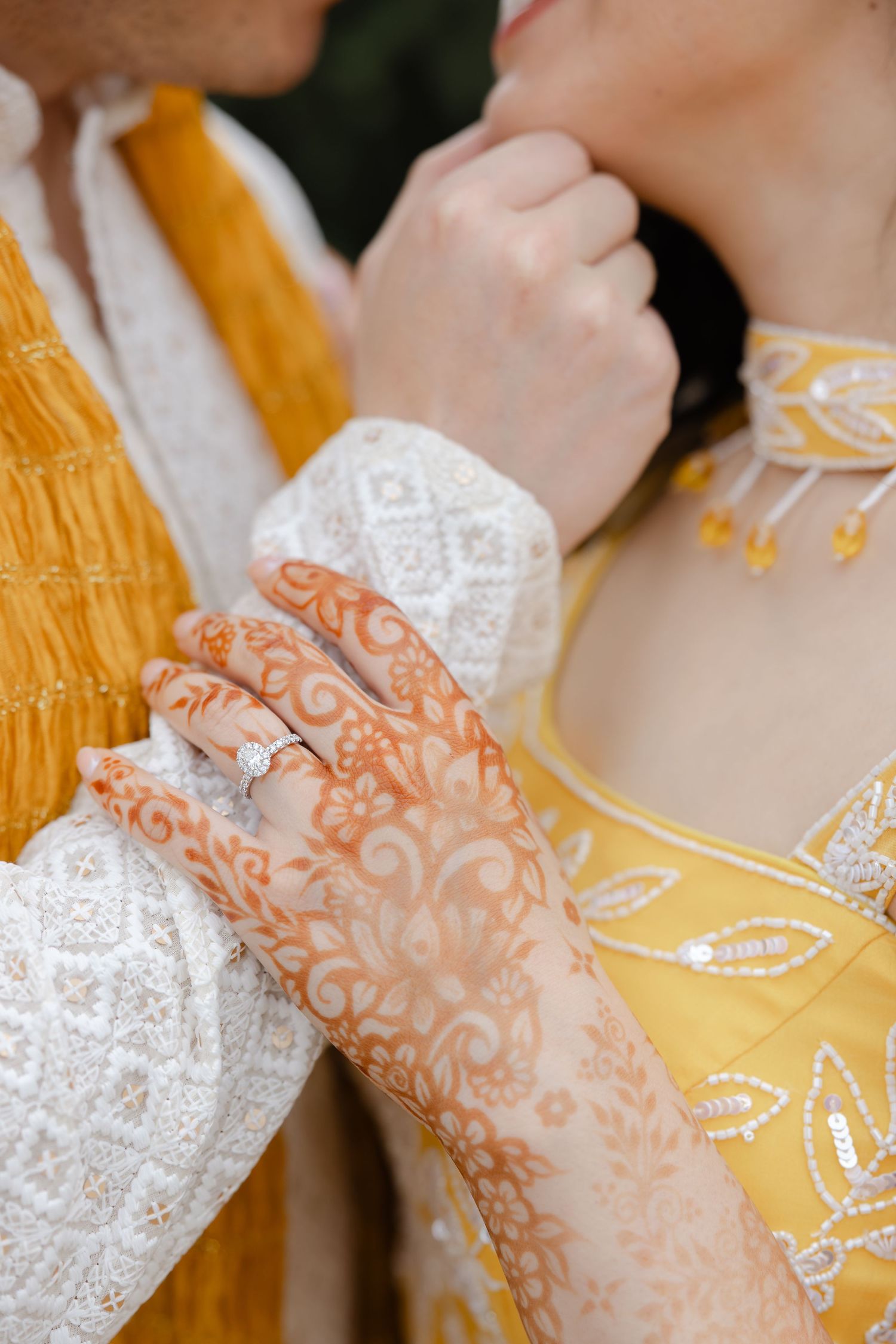 Close-up of henna-adorned hands with diamond ring, yellow embroidered bridal outfit, and white lace sleeve.
