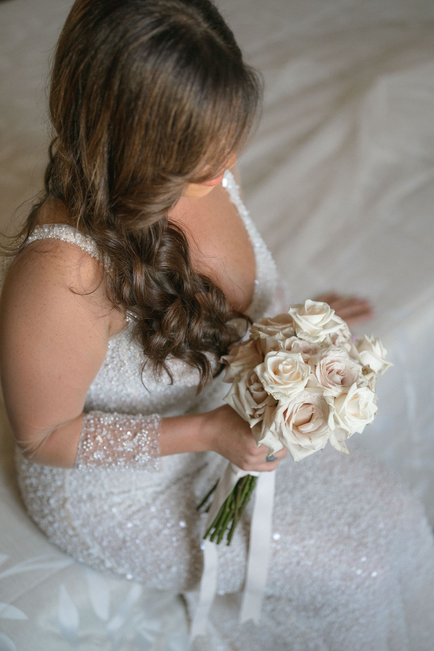 A bride in a lace wedding dress holds a classic bouquet of white roses while seated.