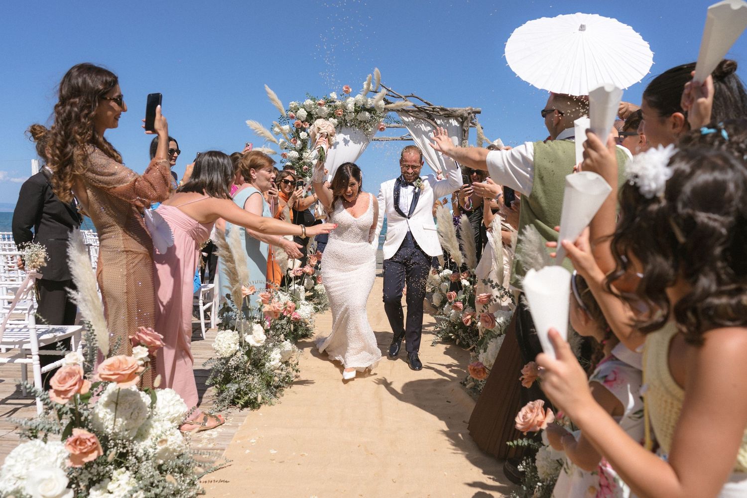 Wedding guests celebrate under a floral arch at a beachside ceremony with parasols.