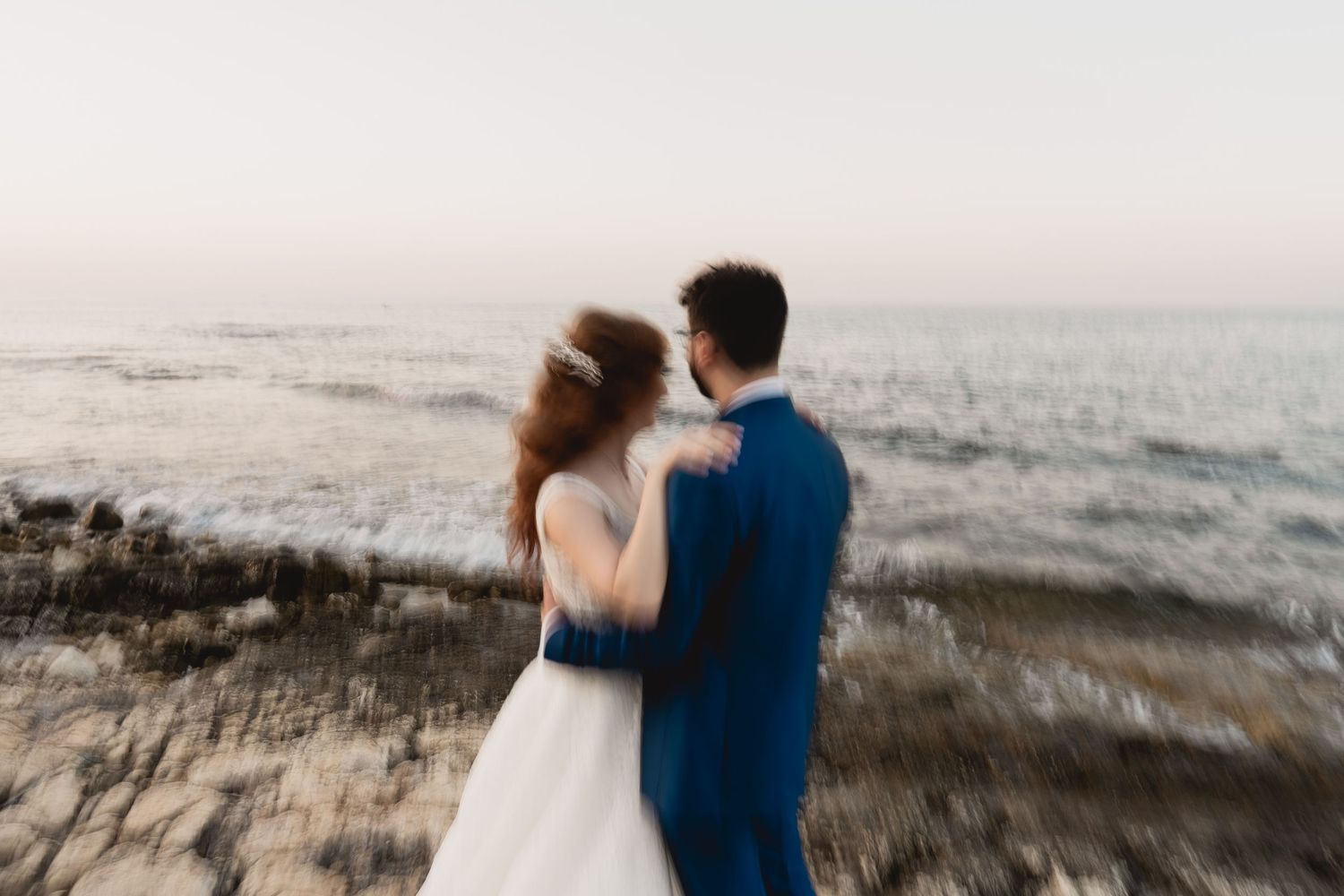 A couple embraces while standing on a sandy beach at sunset with ocean waves in the background.