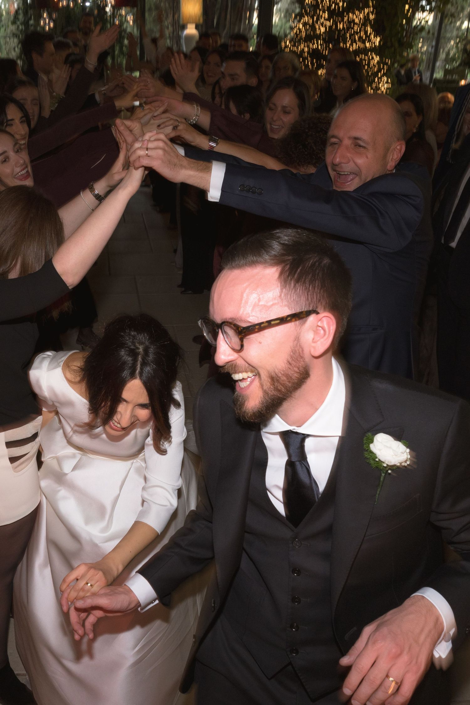 Groom in dark suit laughs as wedding guests celebrate with raised hands on dance floor.