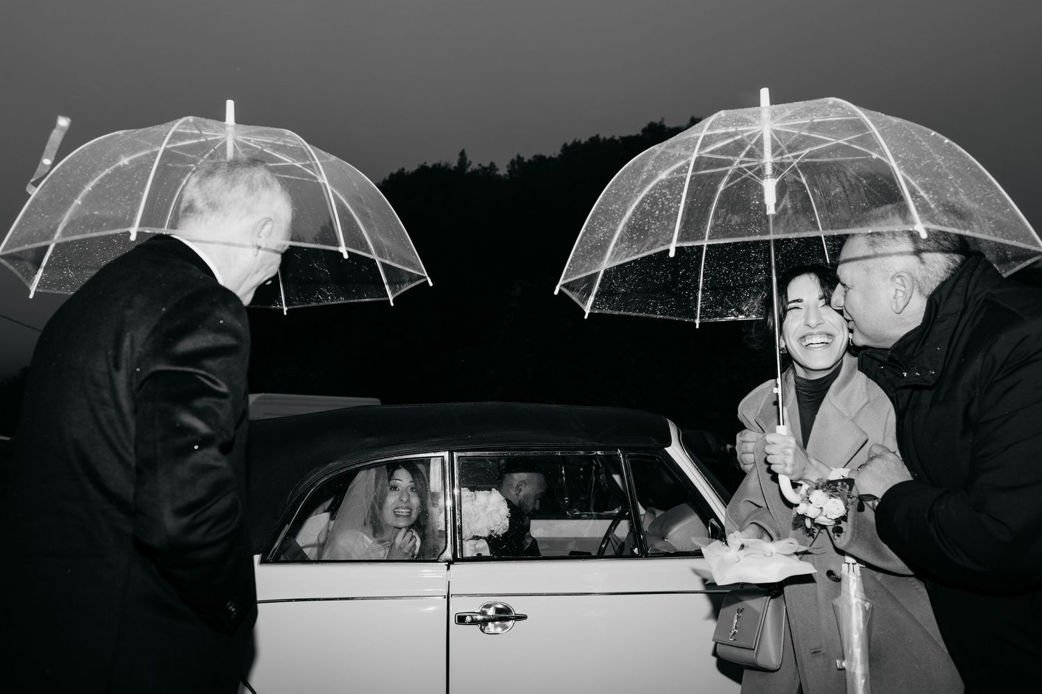 Black and white photo of guests with clear umbrellas greeting a couple arriving by car in the rain.