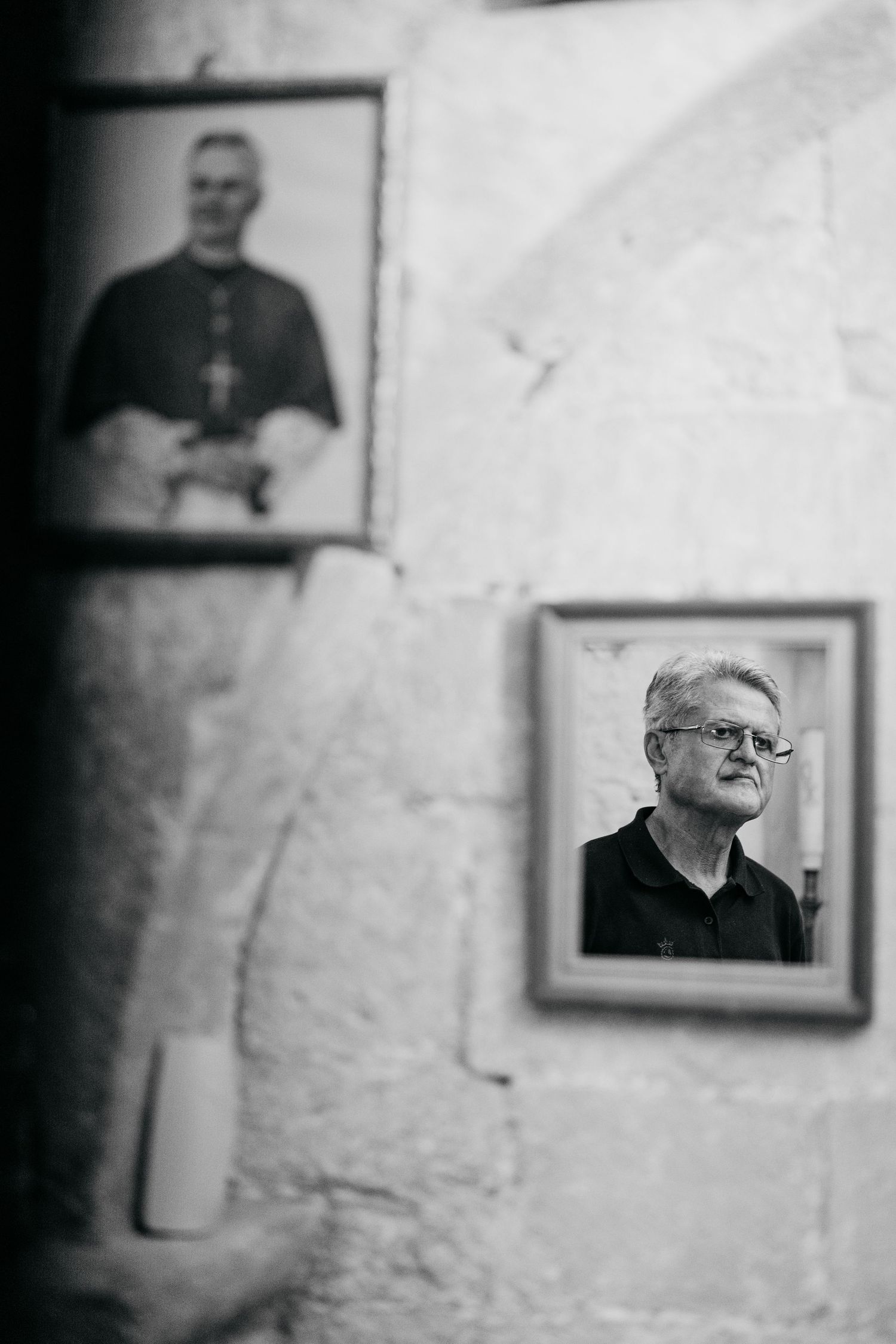 Black and white photo of two framed portraits on a stone wall, one showing an elderly man with glasses, another a clergy member.