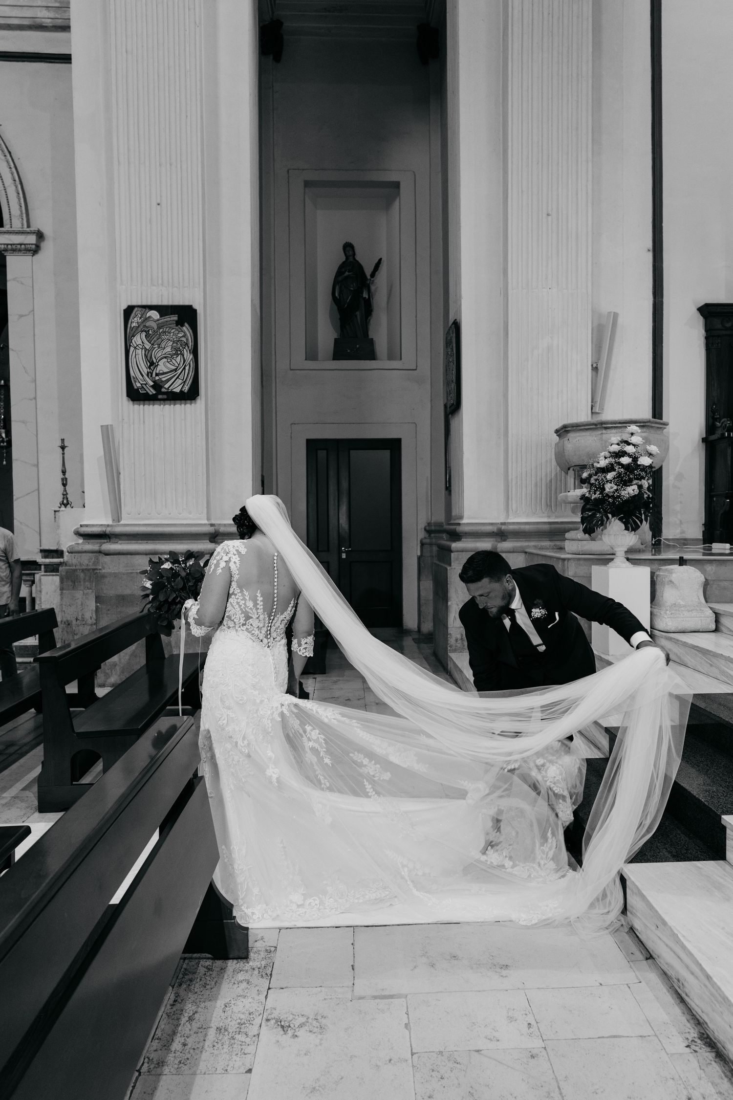 Groom arranges bride's long cathedral veil inside elegant church with classical columns and religious statues.