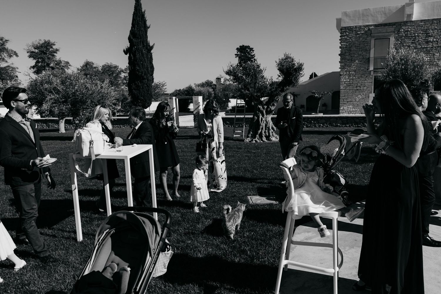 Black and white photo of guests mingling outdoors at a garden wedding reception with white high tables.