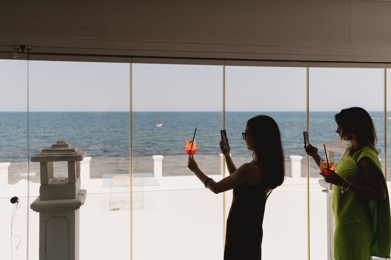 Two silhouettes stand against large windows overlooking a beach while holding drinks at sunset.