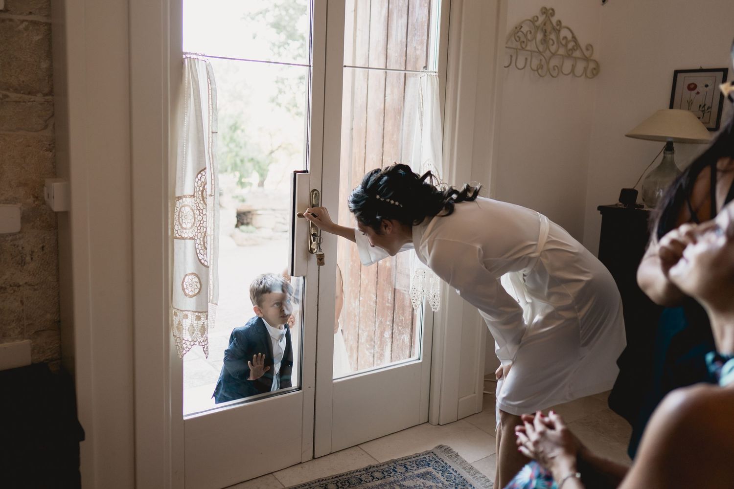 Through a glass door, a child peeks in from outside as someone in white leans to look back through the window.