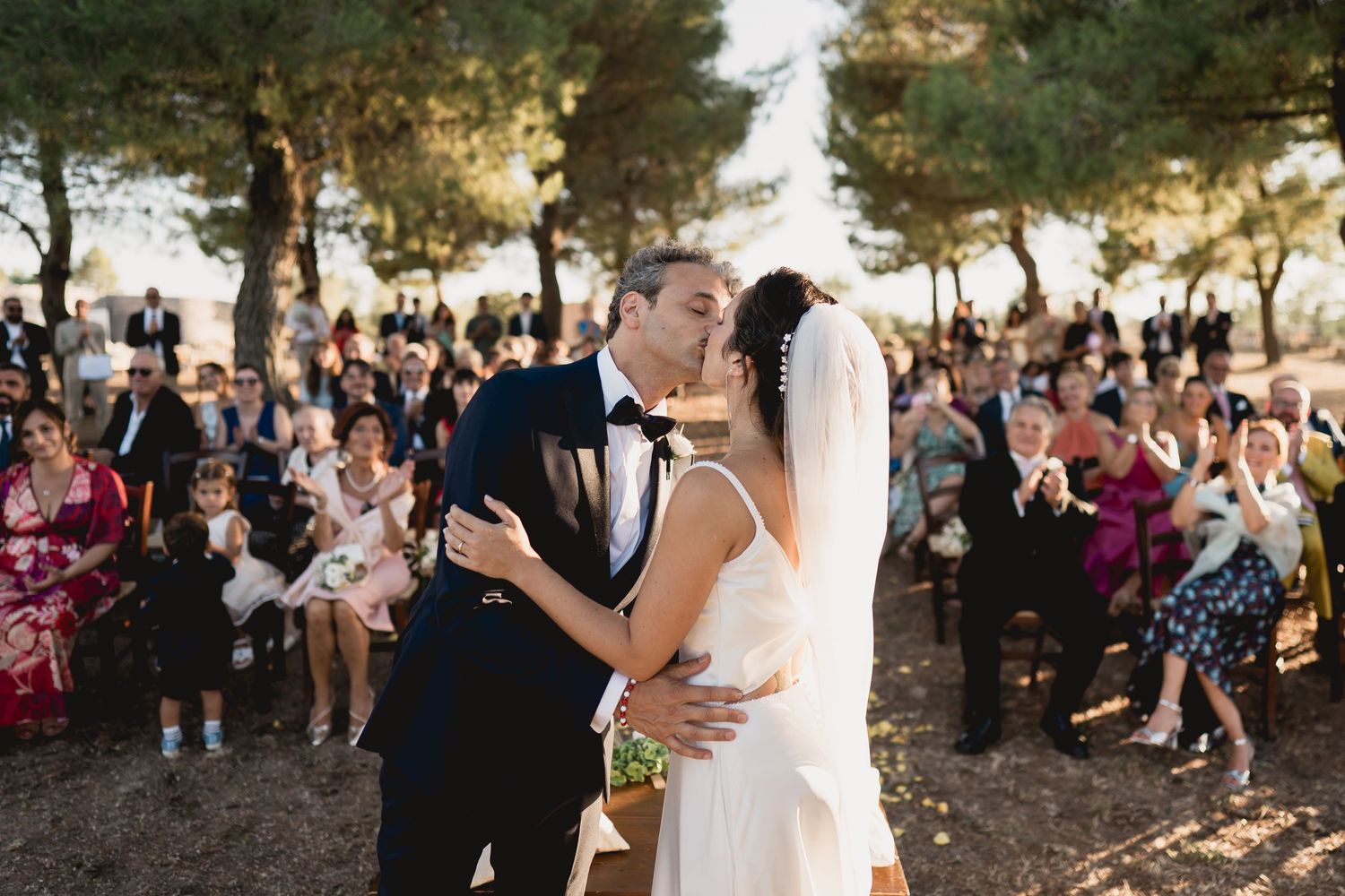 A romantic outdoor wedding ceremony with guests watching the couple share their first kiss under pine trees.