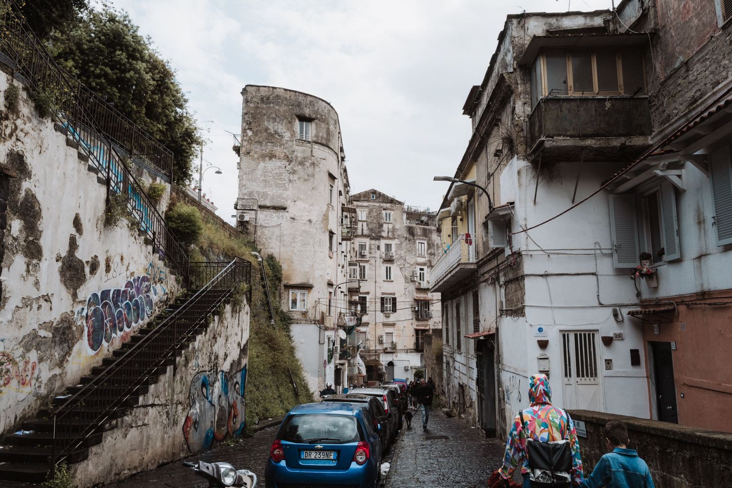 Narrow street winds through old weathered buildings with stairs and graffiti in an urban neighborhood.