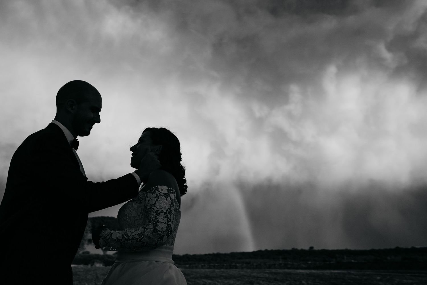 Silhouetted wedding couple embraces against stormy dramatic clouds at sunset.