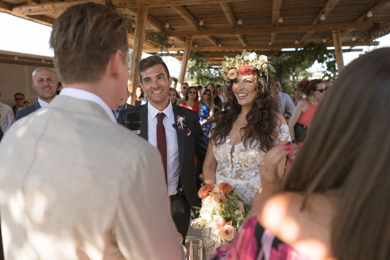 Wedding guests mingle and socialize under a covered outdoor venue decorated with floral arrangements.