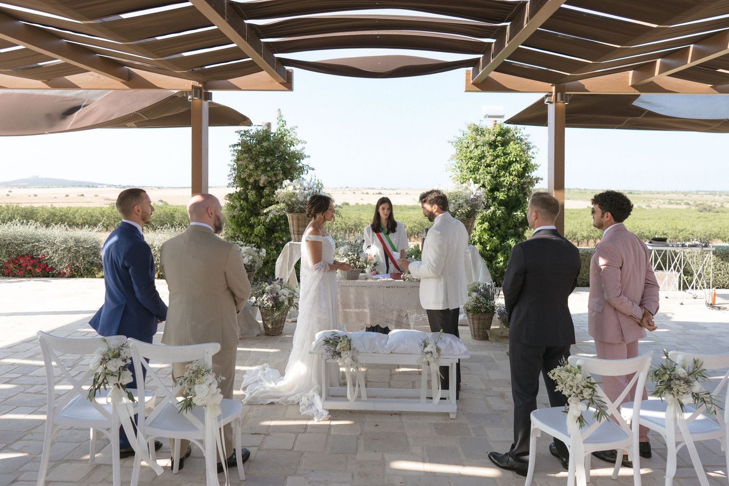 Wedding ceremony setup with white chairs and wooden pergola covering in an outdoor desert setting.