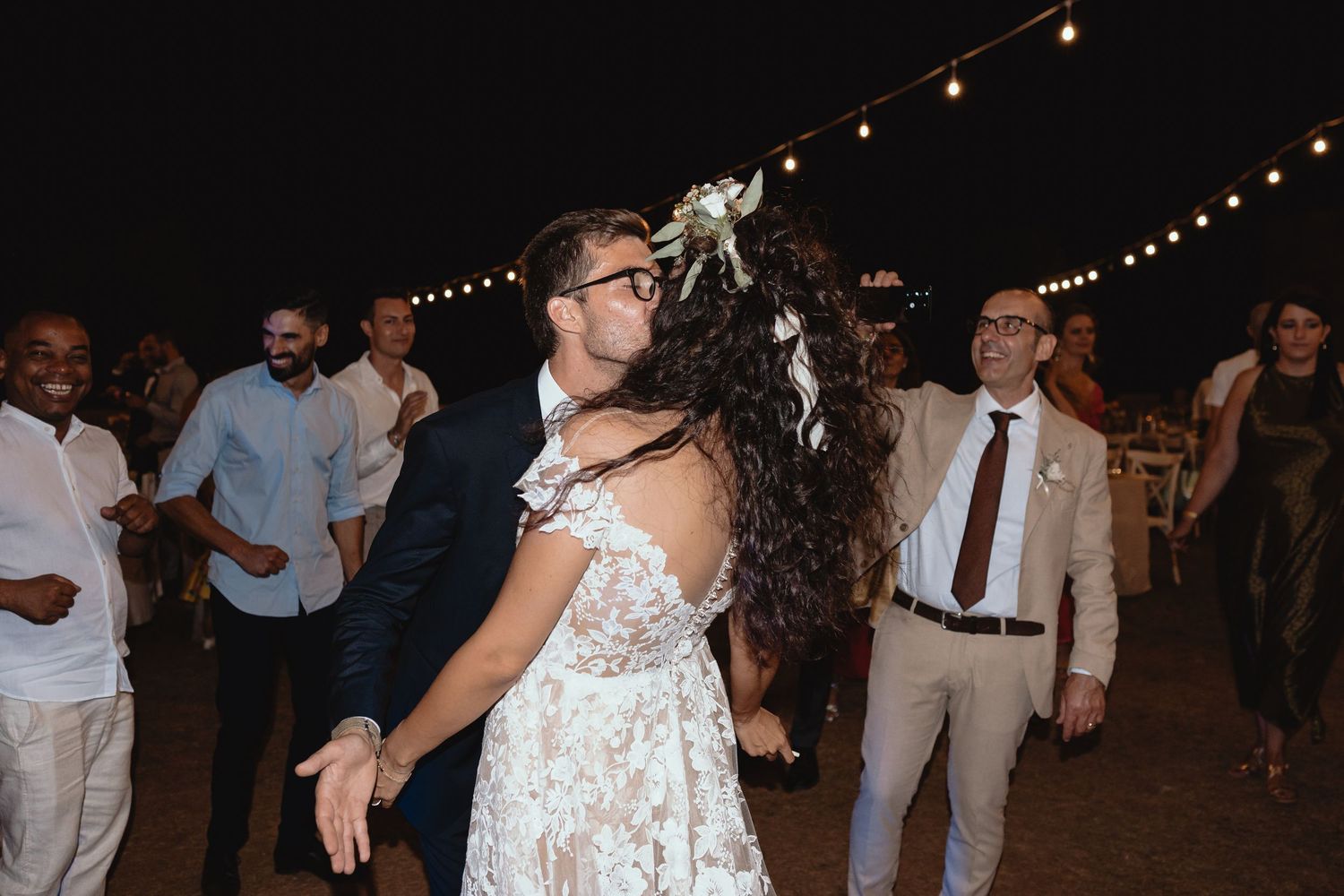 Wedding guests dancing and celebrating at an outdoor evening reception illuminated by overhead string lights.