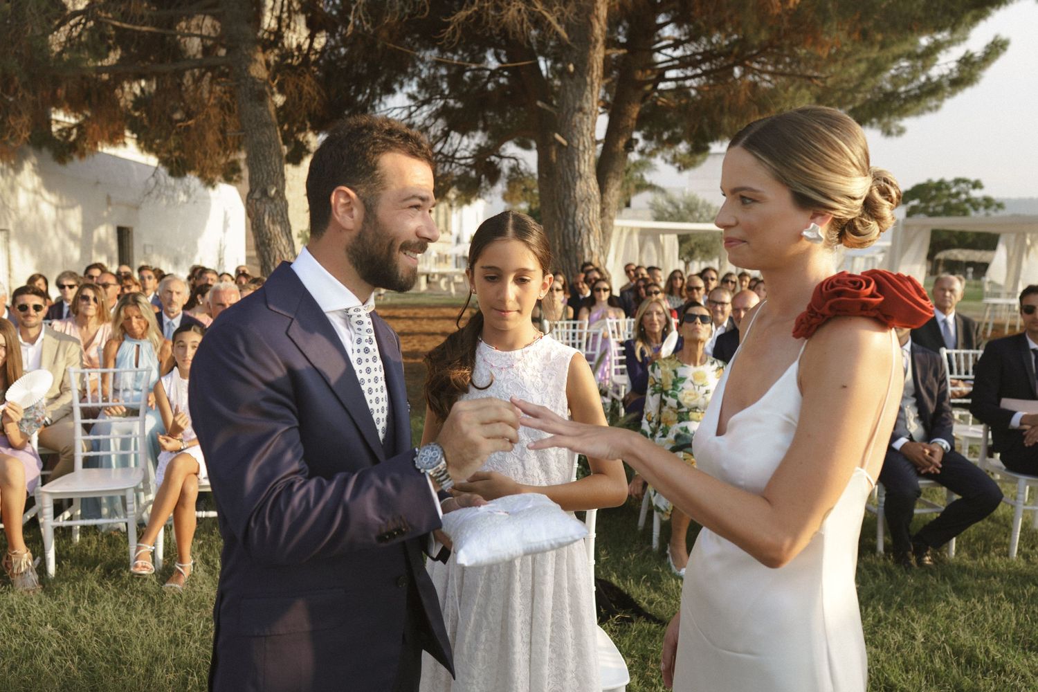 An outdoor wedding ceremony taking place under pine trees with white chairs set up on a lawn.