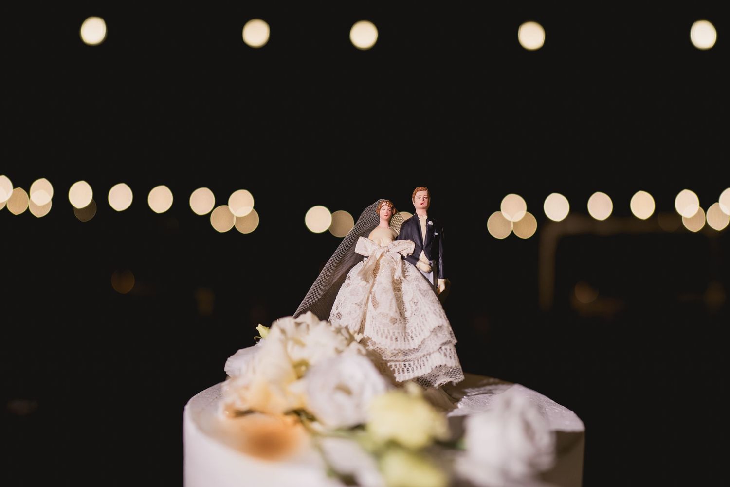 Miniature bride and groom cake topper on white wedding cake with string lights in background.