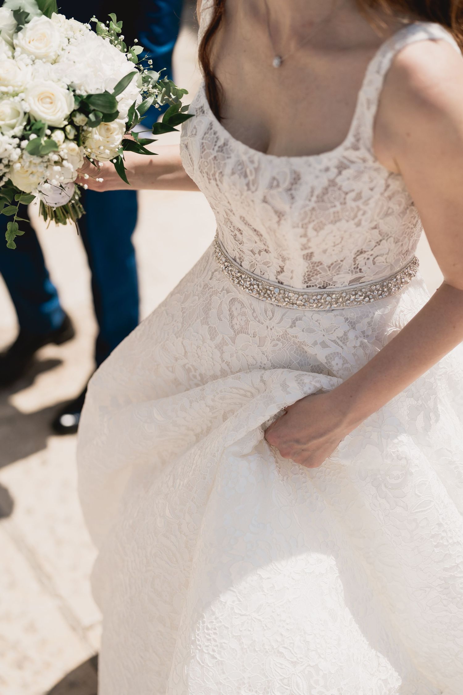 Elegant lace wedding dress with beaded waistband and white floral bouquet in natural sunlight.