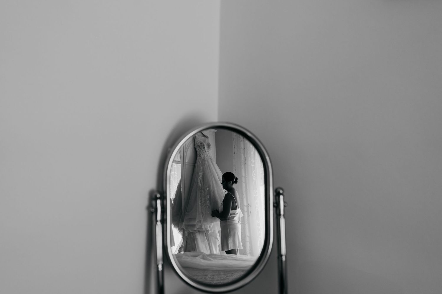 Wedding dress reflection in an oval vanity mirror against grey wall.