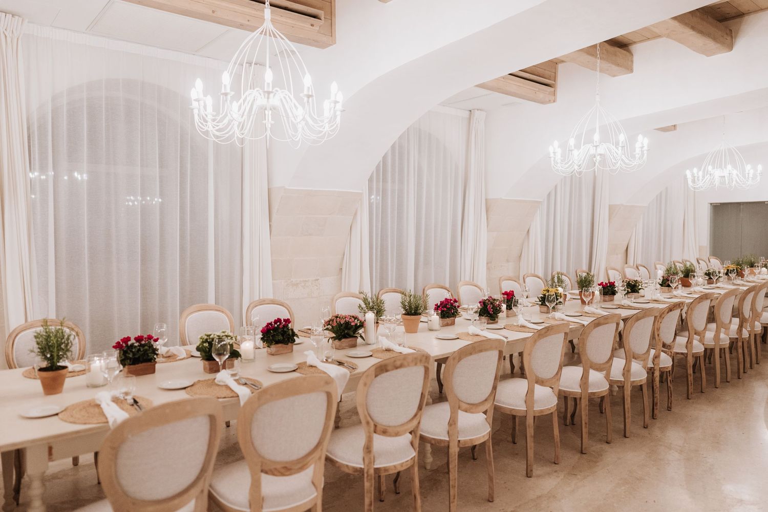 Elegant wedding reception table setup with white chairs and red floral centerpieces under crystal chandeliers.