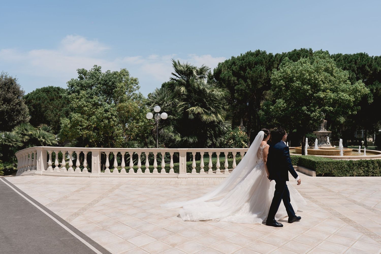 A couple walks along an elegant stone balustrade walkway with the bride's veil trailing behind.