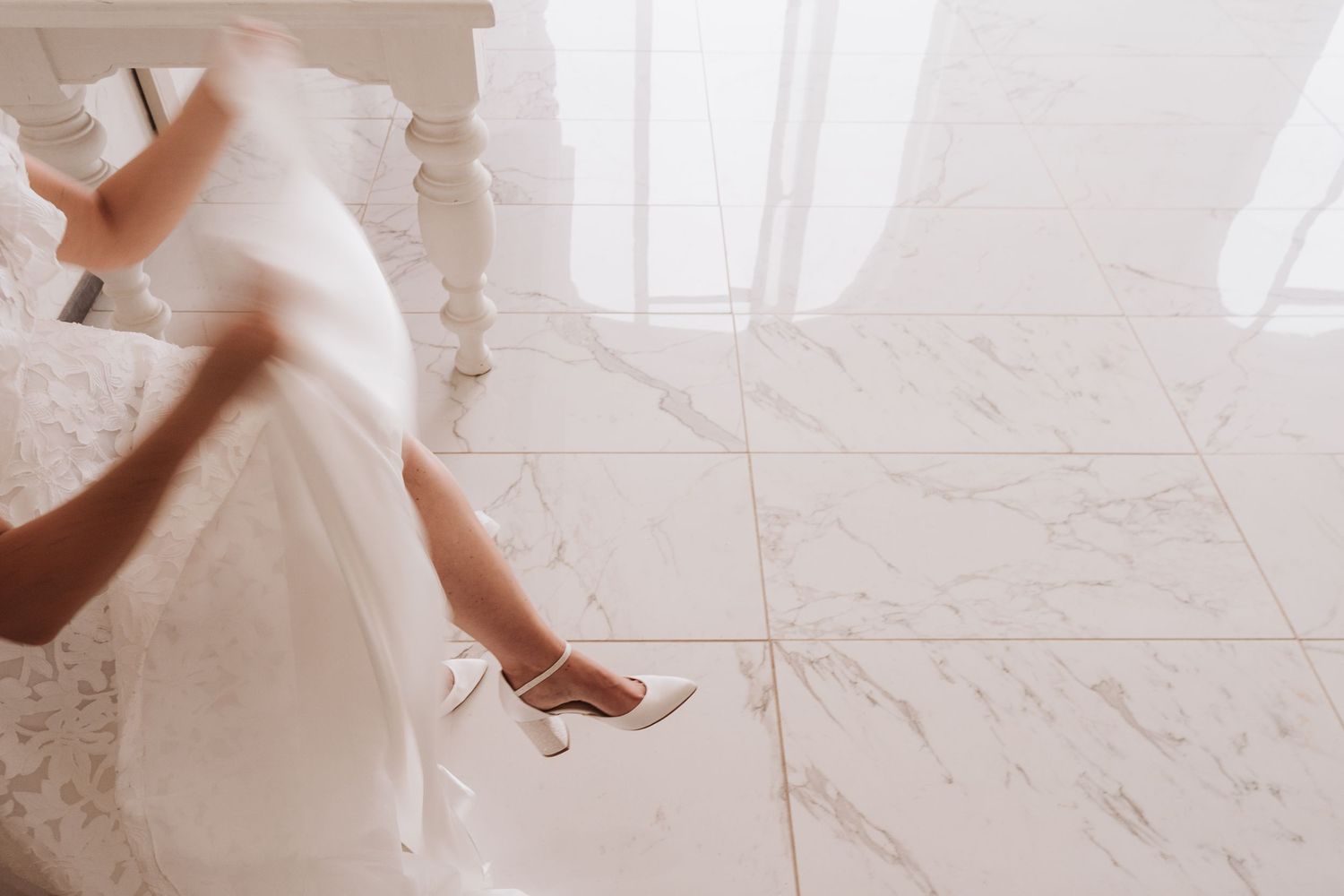 Close-up view of flowing white wedding dress and high heels against marble floor.