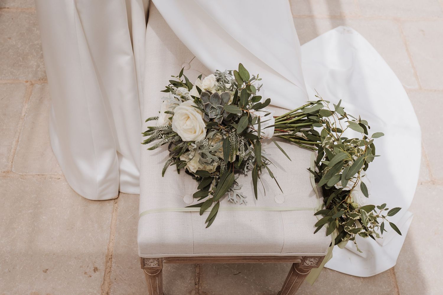 Two elegant white wedding bouquets with greenery resting on a light-colored chair.