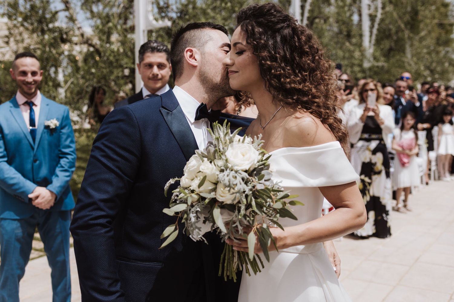 Couple shares intimate moment during outdoor wedding ceremony with white floral bouquet.