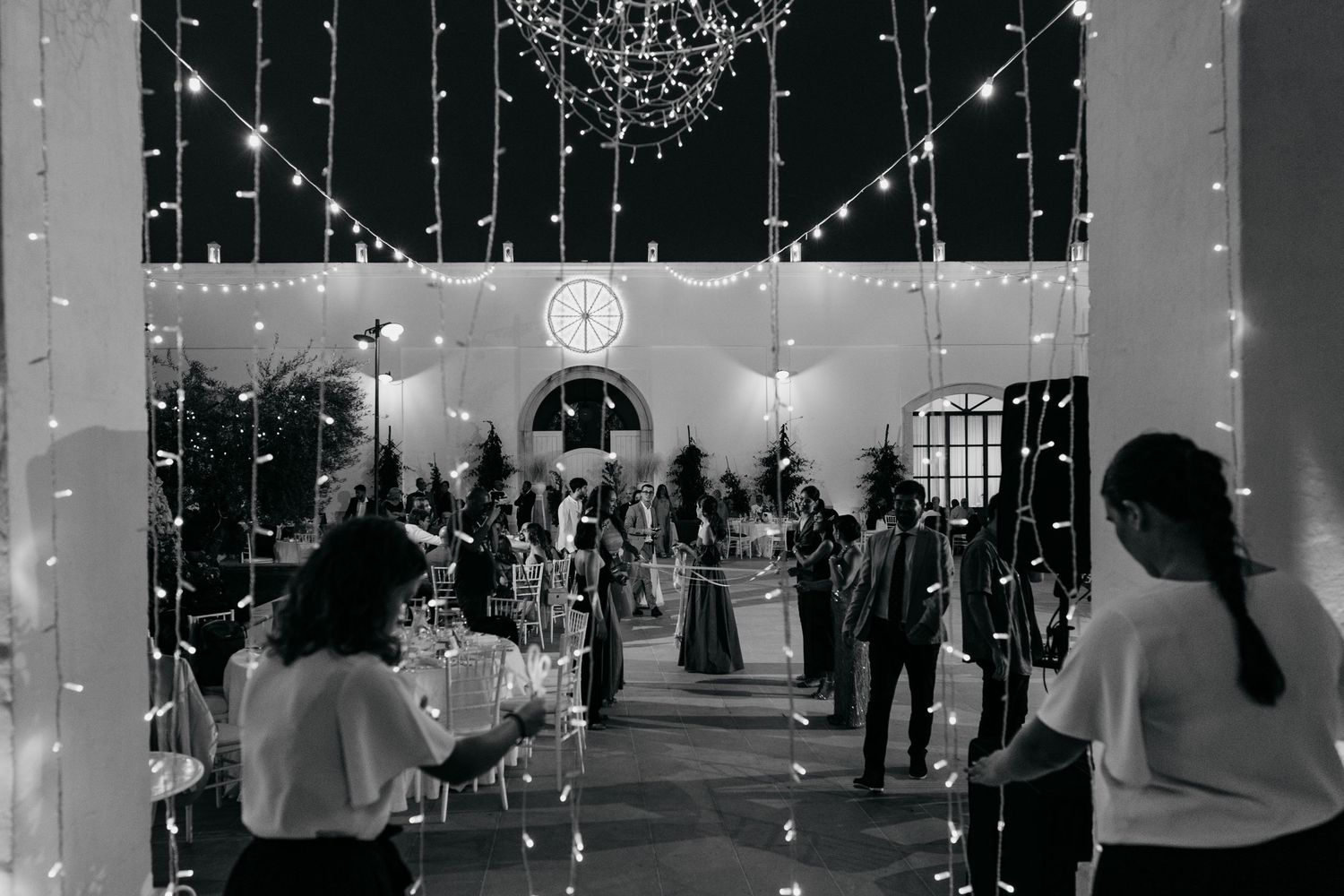 Wedding guests gather under twinkling string lights in an elegant outdoor venue for an evening celebration.