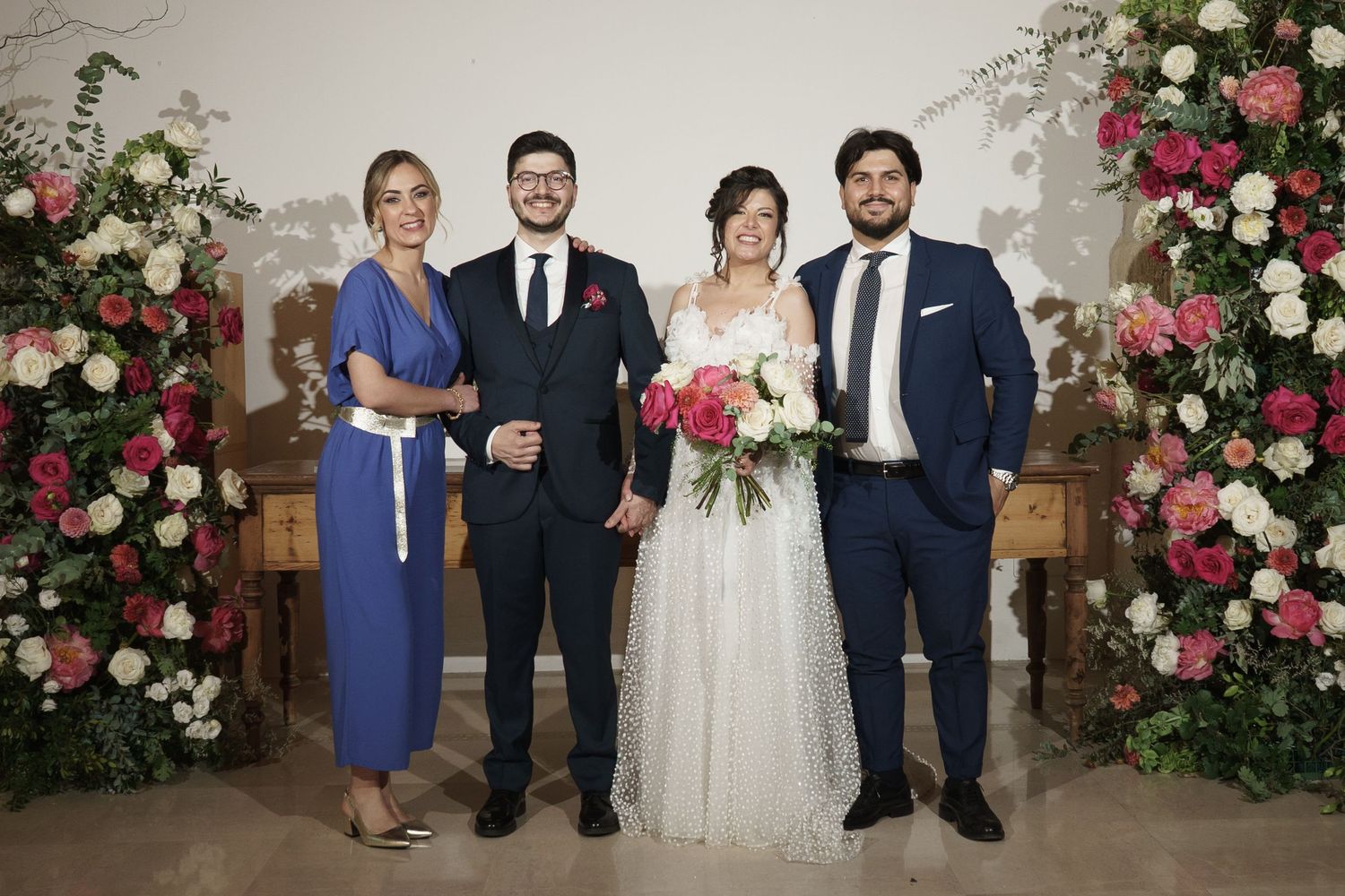 Wedding party of four posing with floral arrangements, bride in white gown holding bouquet, groom in suit, guests in blue attire.