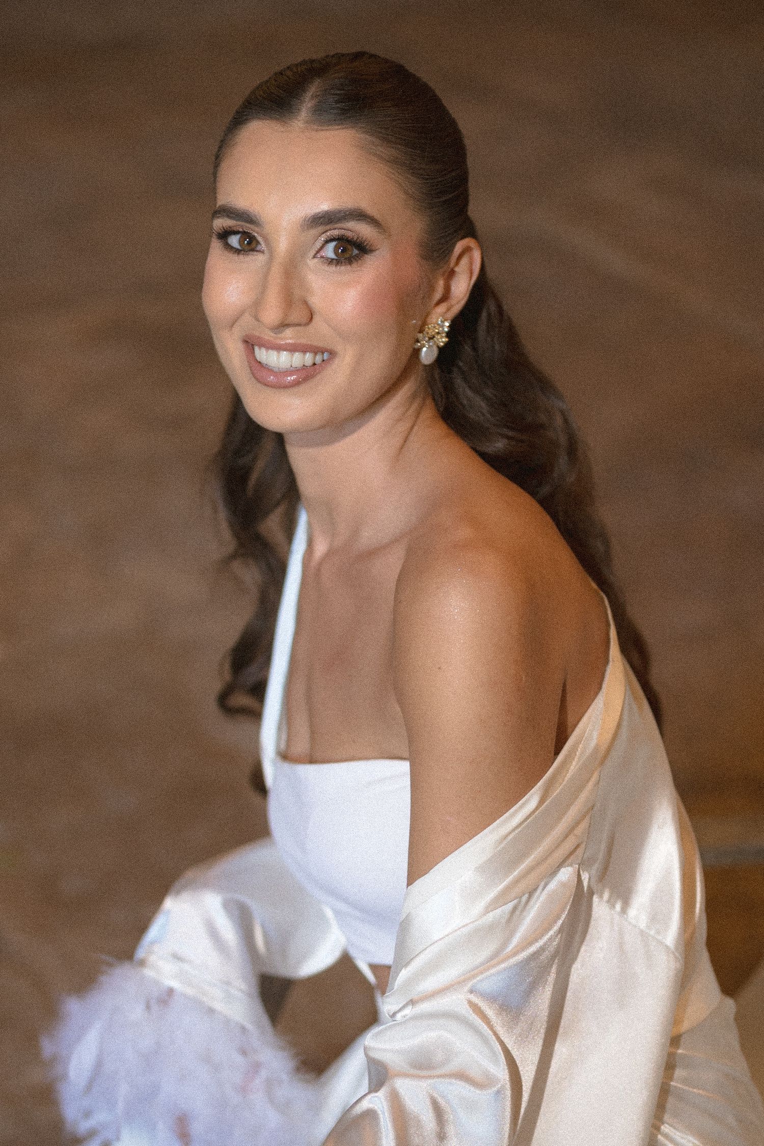 Smiling woman with long dark hair in a low ponytail, wearing a white strapless top, satin robe, floral earrings and a feathered accessory.