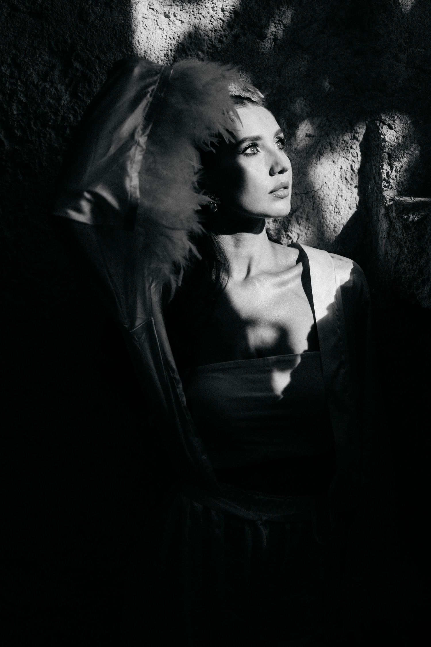 Black and white portrait of a woman in a hooded jacket gazing upward, her face lit by dramatic shafts of light against a dark stone wall.