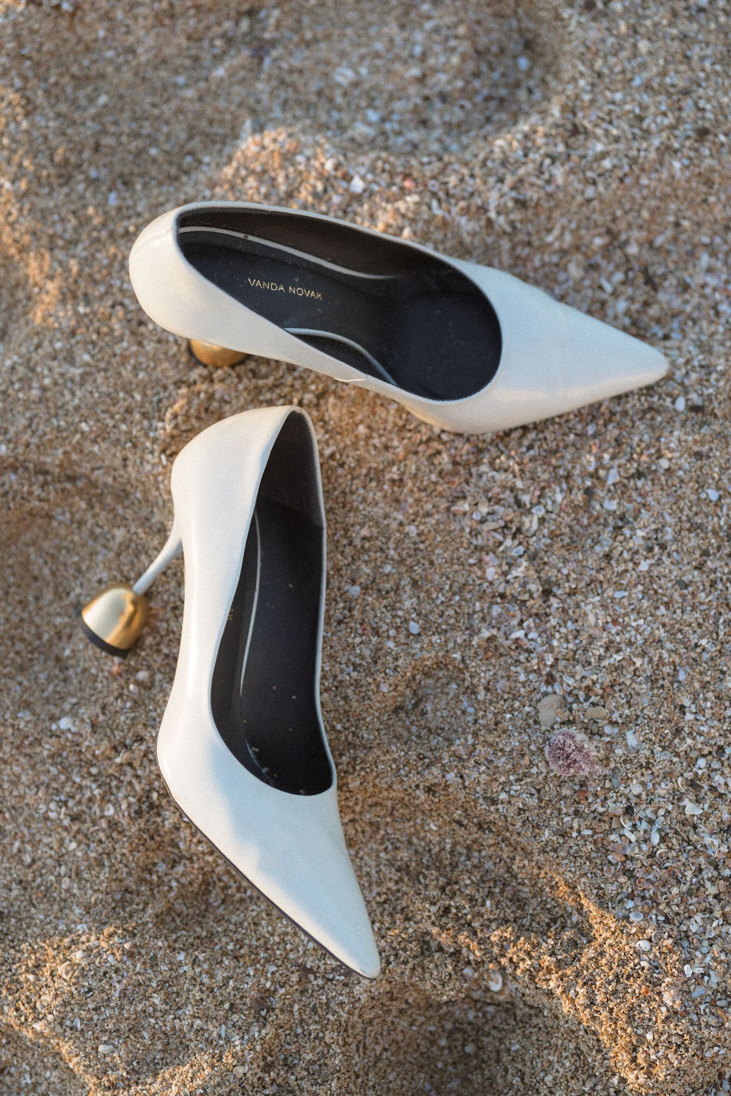 Pair of white pointed-toe bridal heels with gold sculptural heels resting on sandy ground.