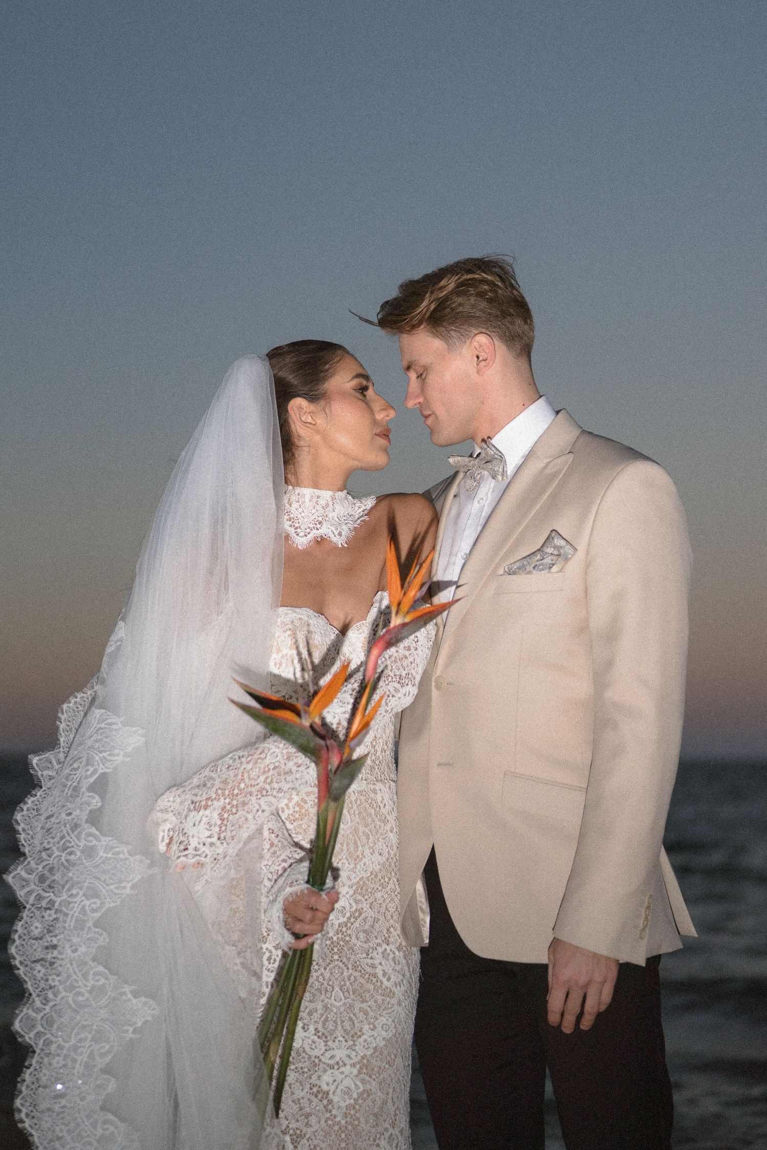 Bride and groom in a beige jacket almost kiss at sunset by the sea, holding a bird of paradise bouquet.