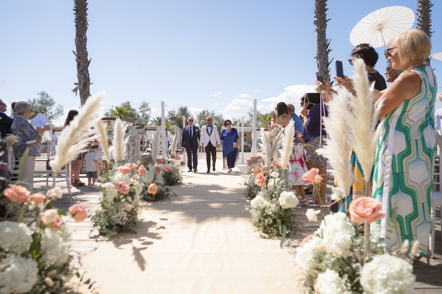 Wedding ceremony aisle decorated with white and peach flowers along a palm tree lined pathway.
