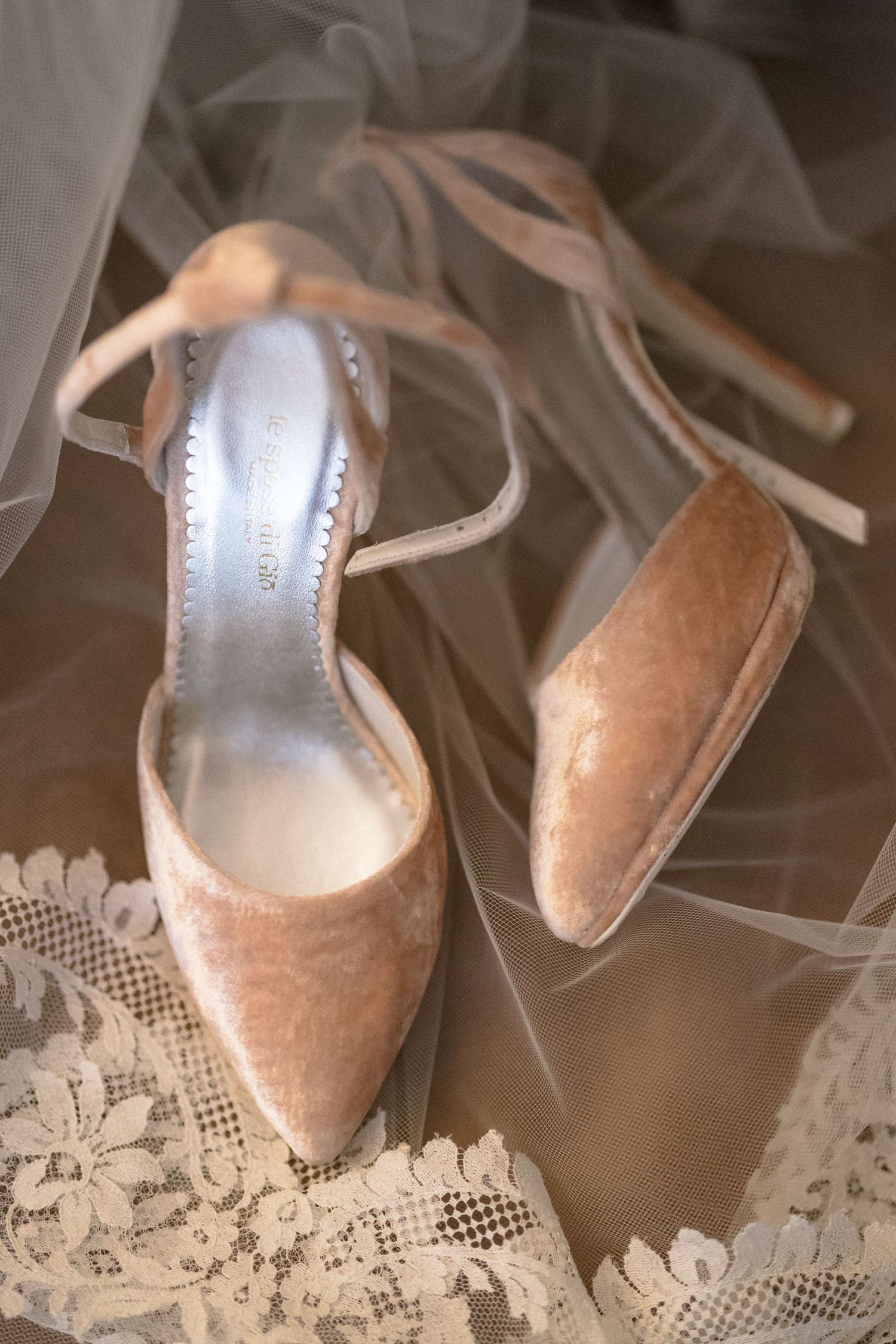 Elegant blush velvet wedding heels with ankle tie straps resting on delicate lace and tulle fabric.