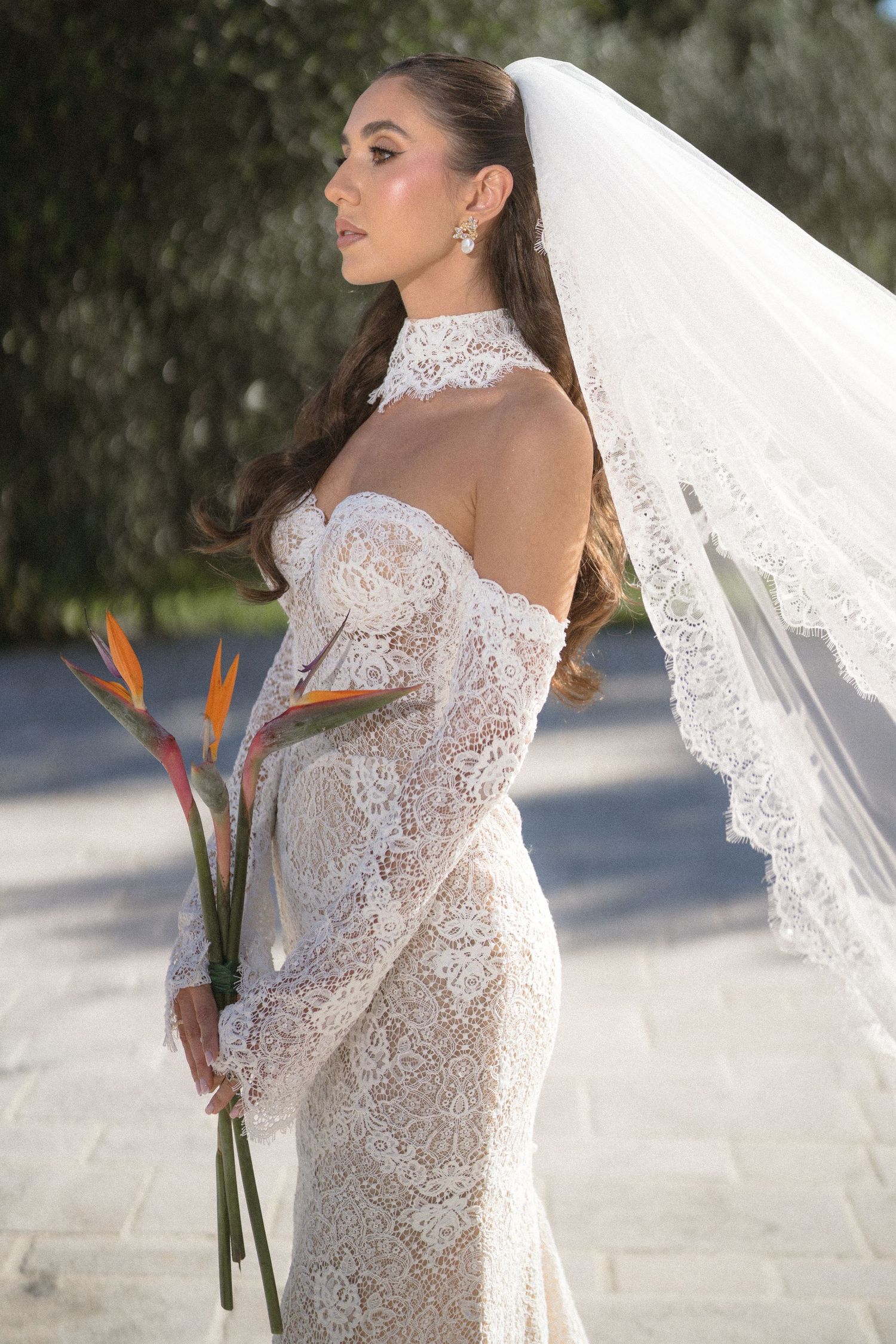Profile of a bride in an off-shoulder lace wedding dress and veil holding a bouquet of orange bird of paradise flowers.
