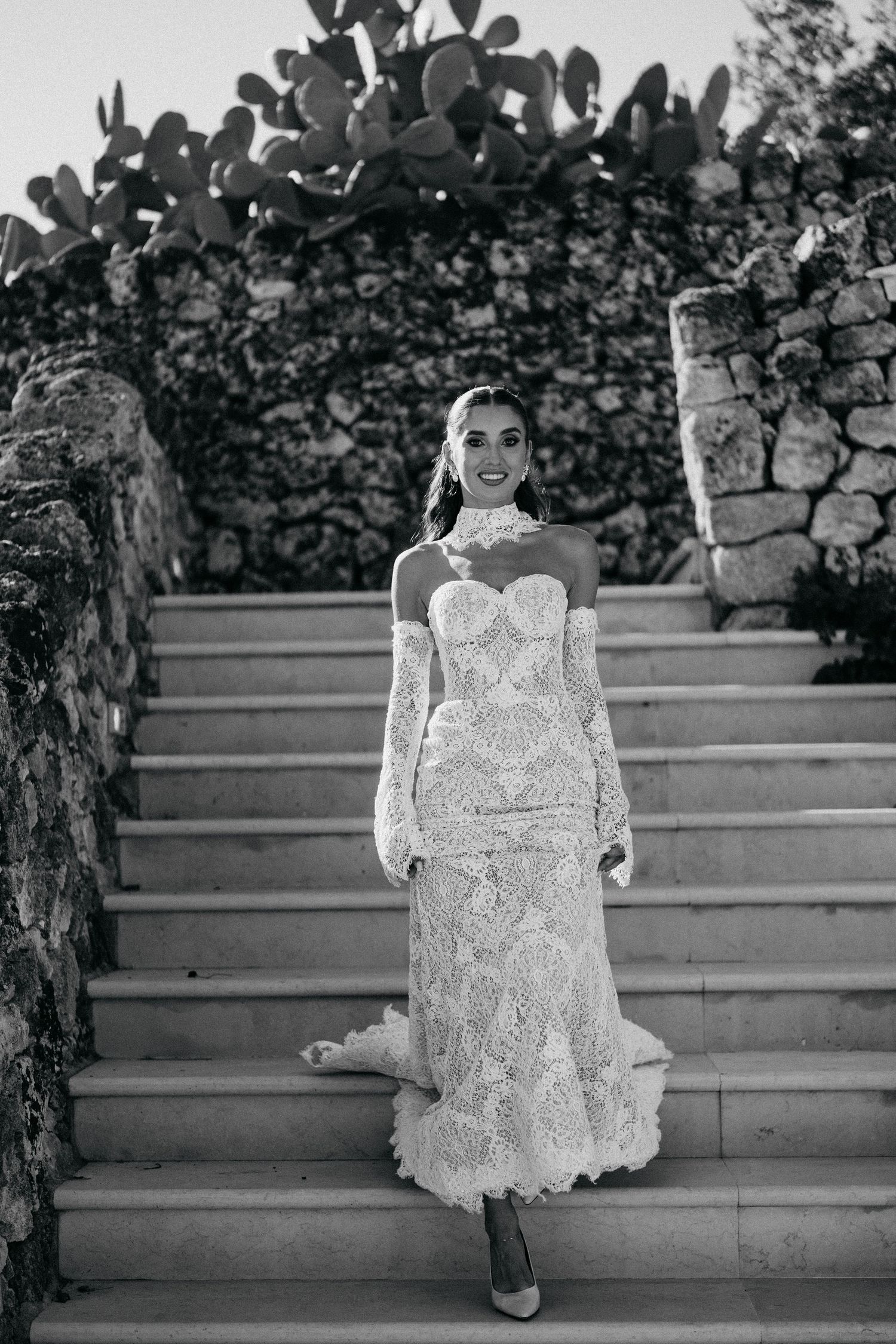 Black and white photo of a bride in a lace gown with a long train descending stone steps beside a cactus-topped wall.