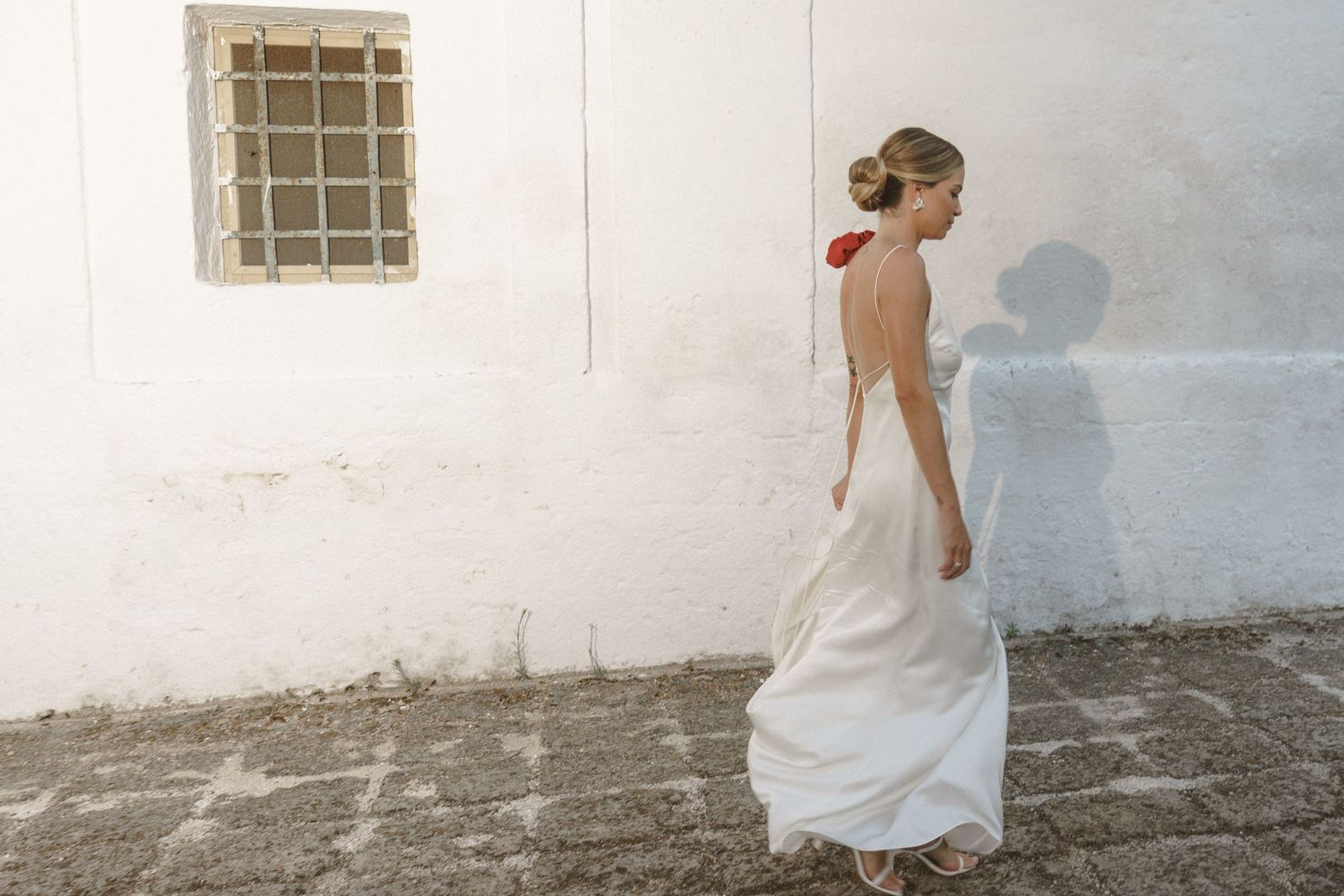 Bride in flowing white dress poses against white Mediterranean-style wall with rustic window.