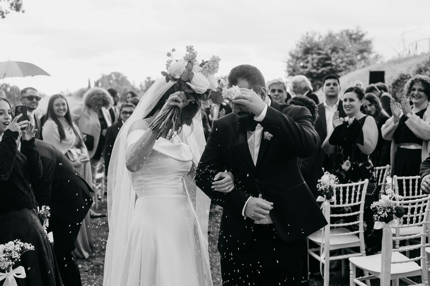 A black and white photo of a joyful wedding celebration with guests cheering at an outdoor ceremony.