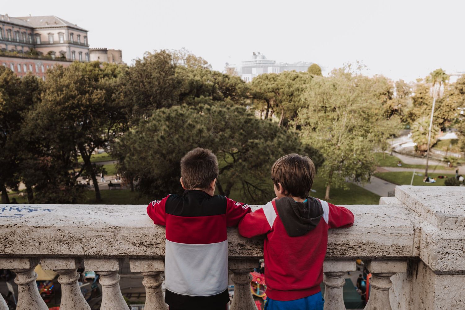 Two children in matching red jackets stand at a stone balustrade overlooking a scenic park view.