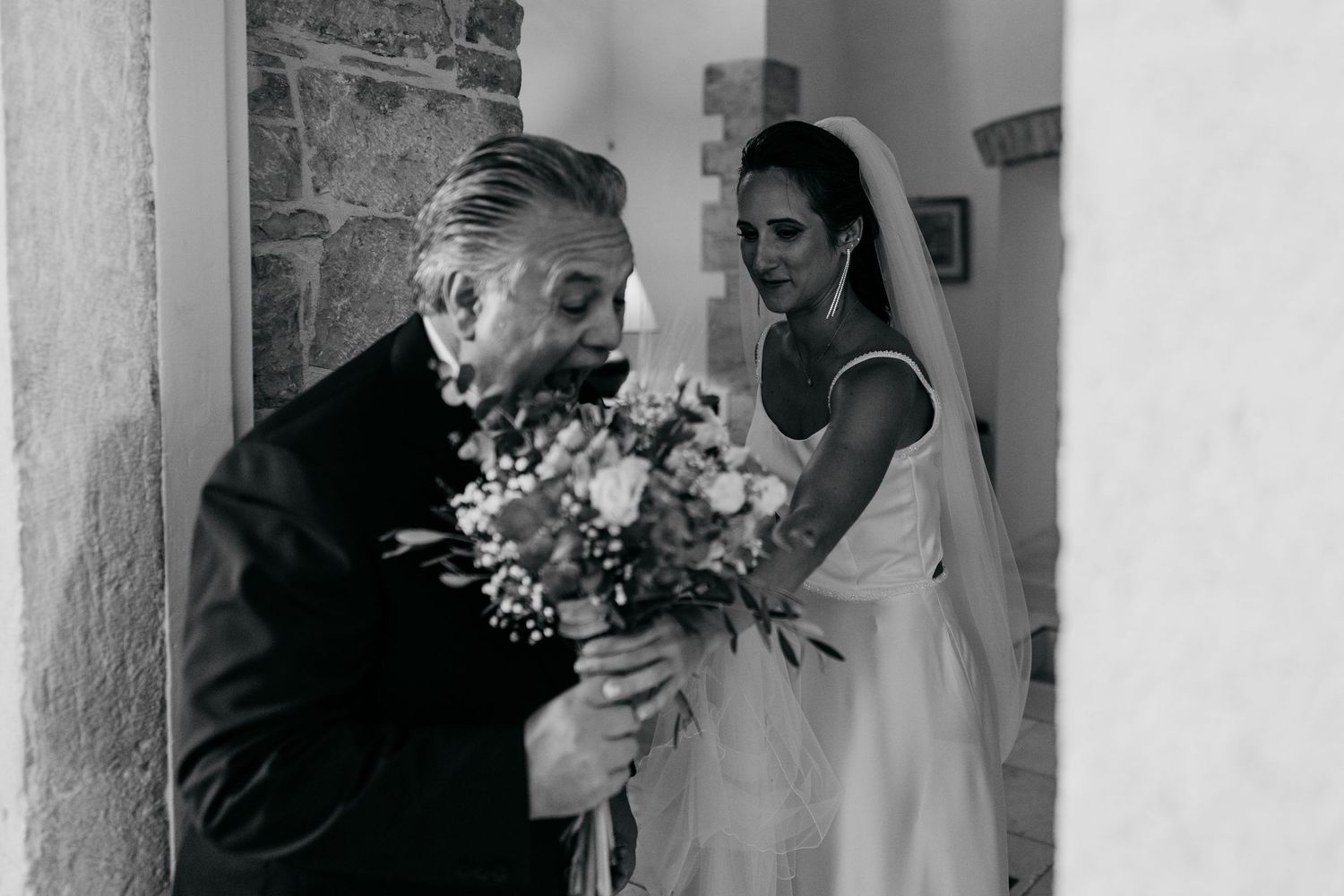 Black and white photo showing joyful wedding scene with flowers in a stone hallway.