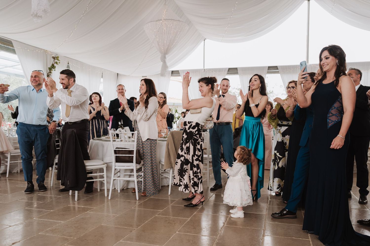 Wedding guests in formal attire standing and applauding during the reception celebration.