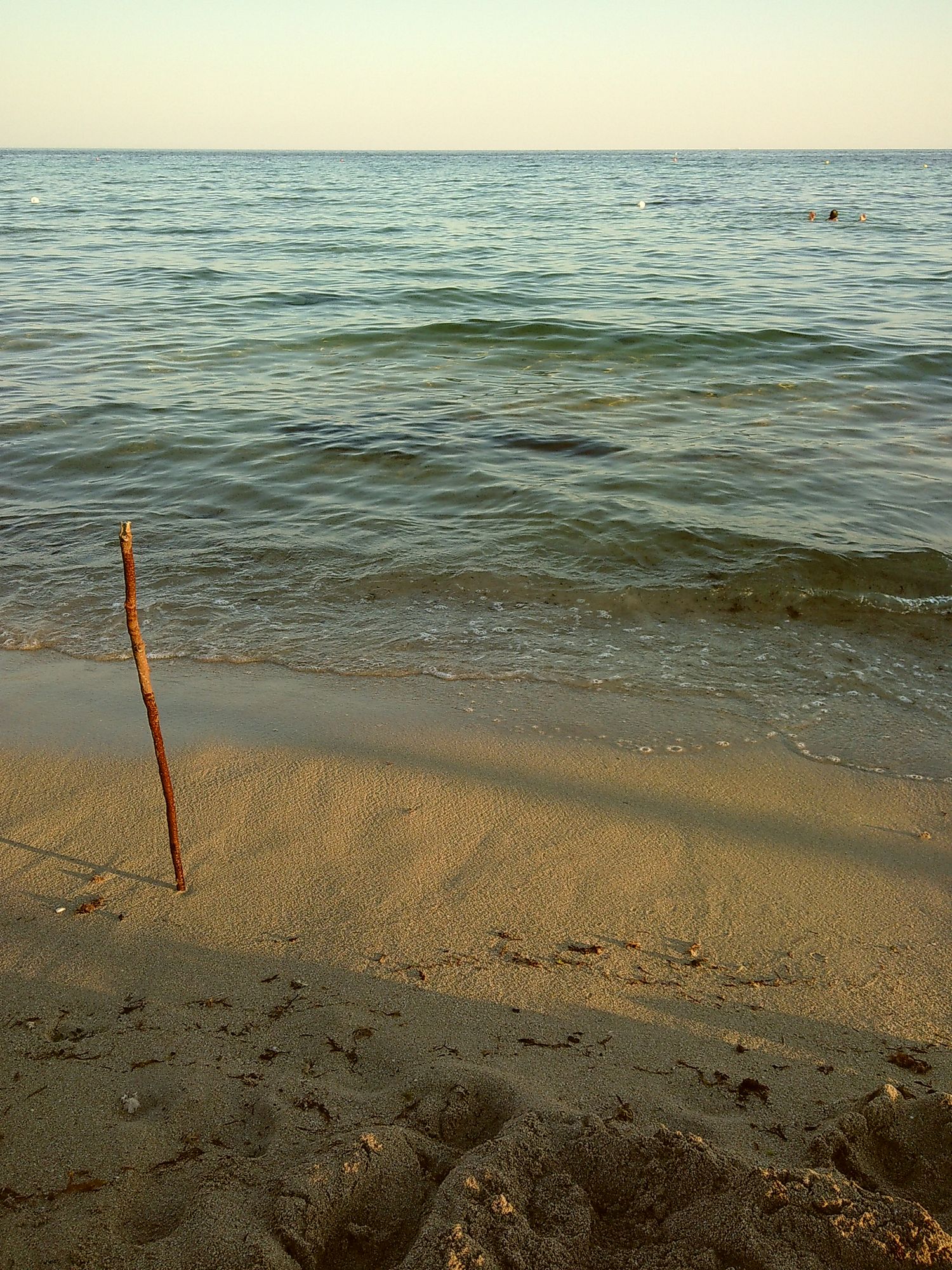 A wooden stake marks the shoreline where gentle waves meet sandy beach at sunset.