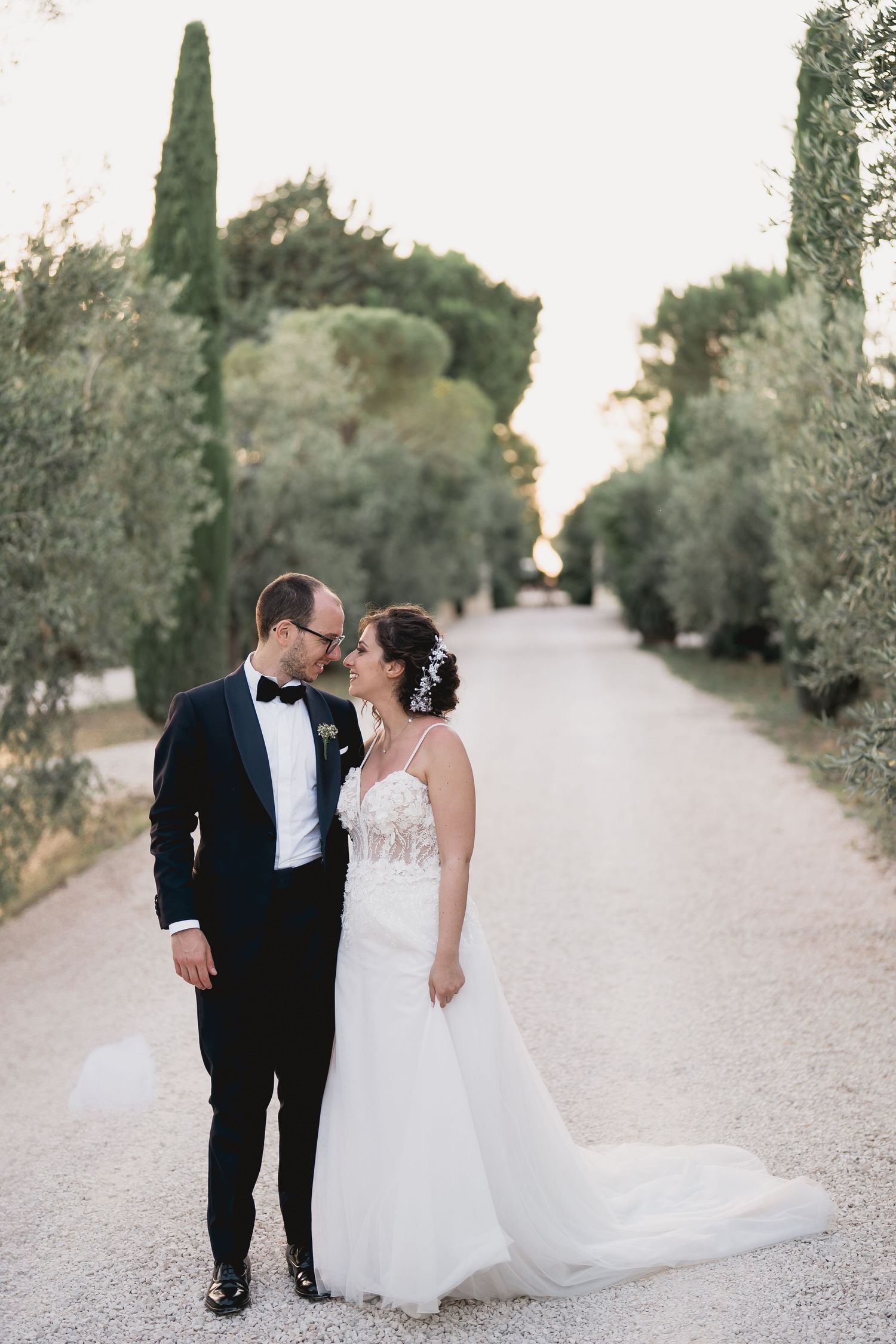 A romantic wedding photo on a tree-lined path with cypress trees and Mediterranean landscaping.