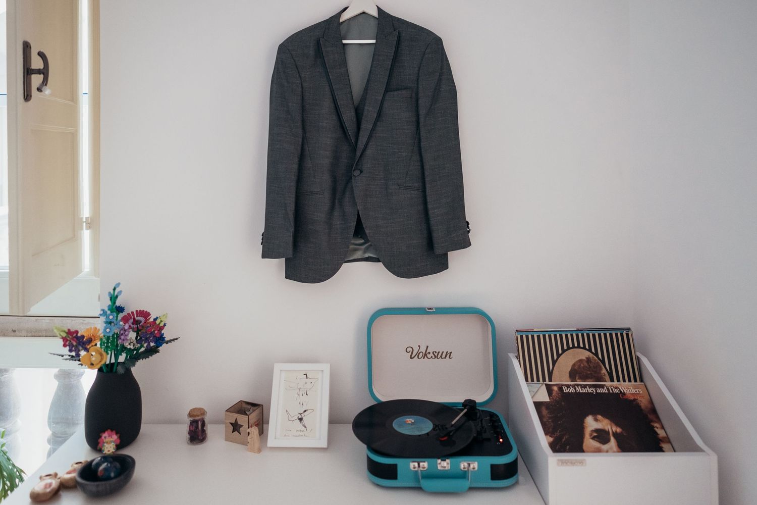 Gray blazer hanging on wall with turquoise record player and decorative items on shelf below.