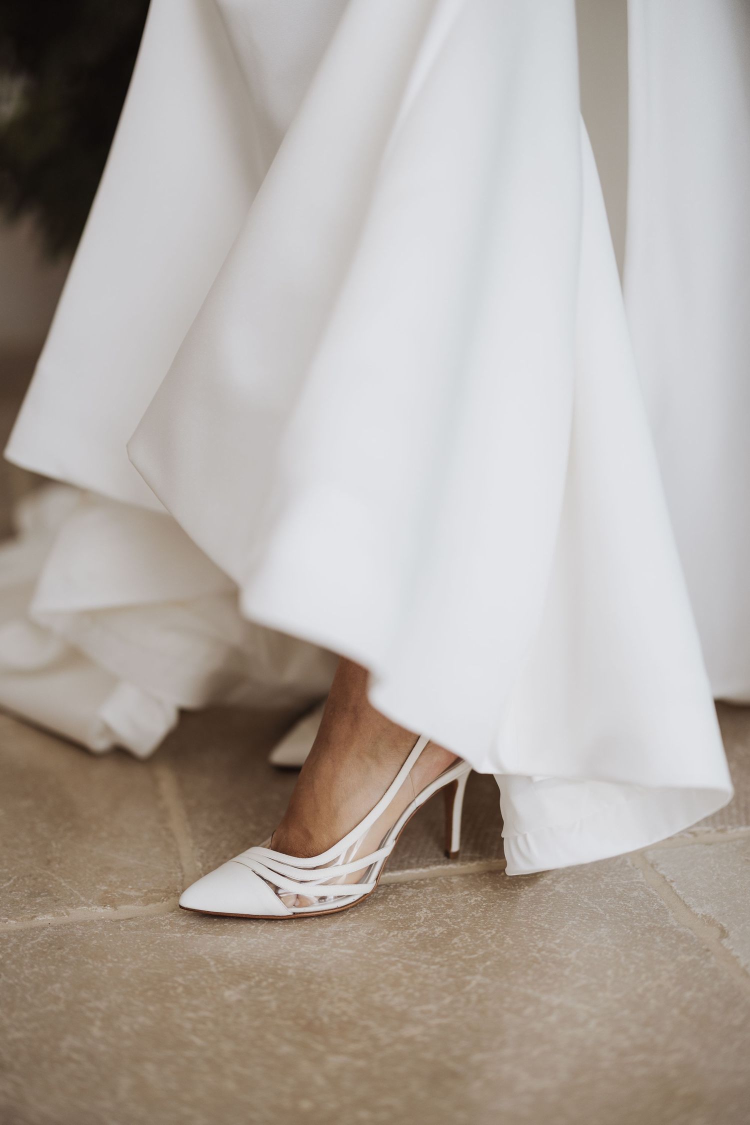 Elegant white bridal shoe peeking out from under flowing wedding dress hem.