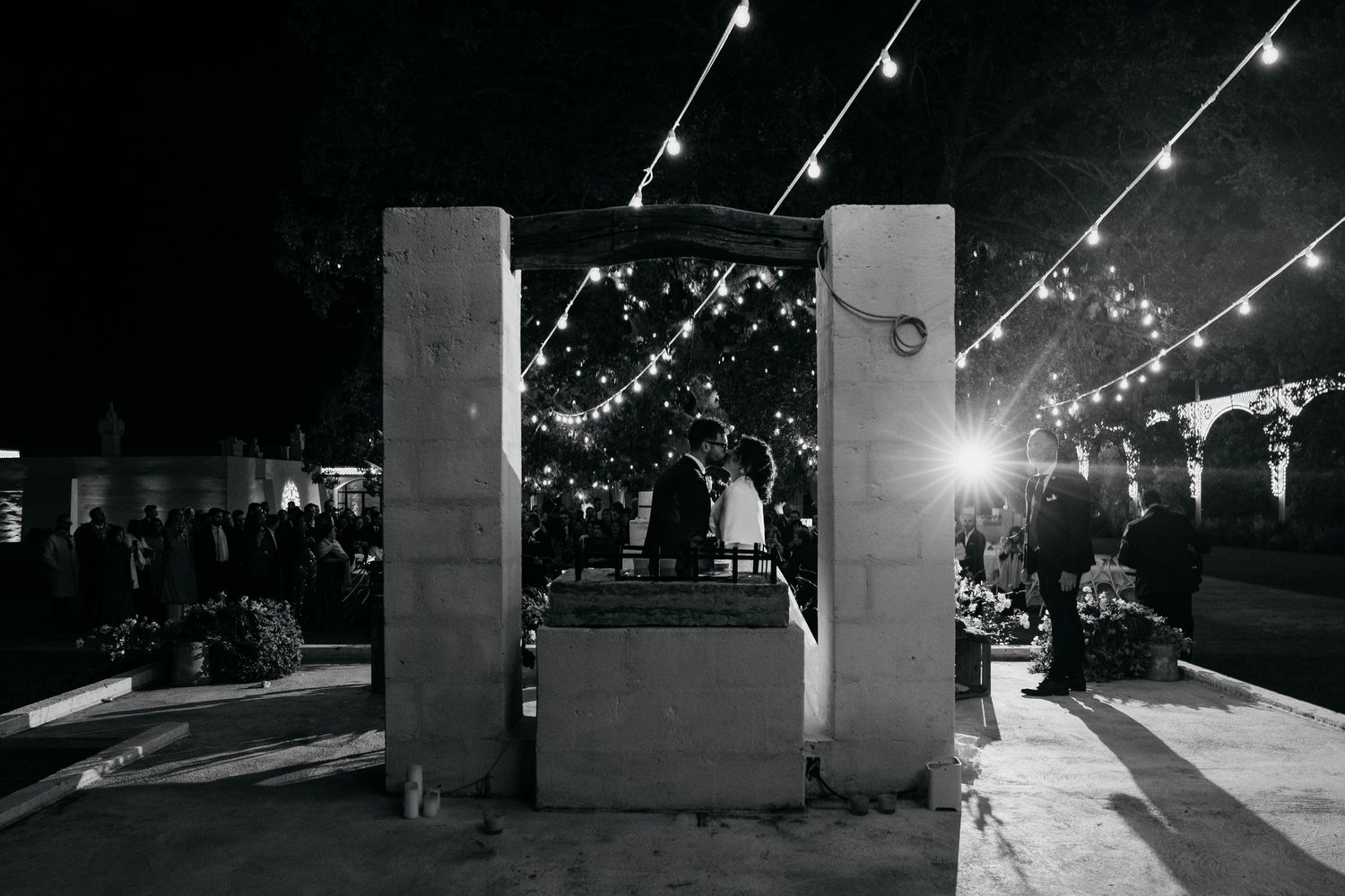Black and white night shot of wedding guests gathered under string lights at outdoor reception venue.